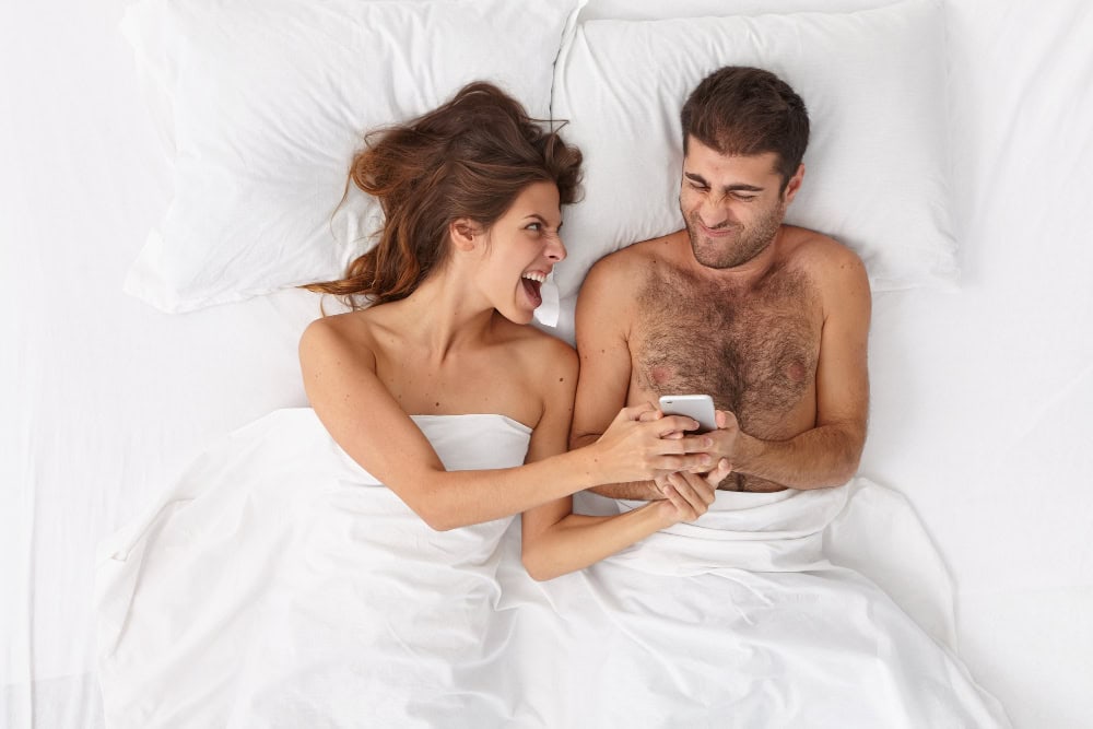 Couple in bed, smiling and playful, symbolizing the use of technology to explore and communicate intimate desires and sexual needs.